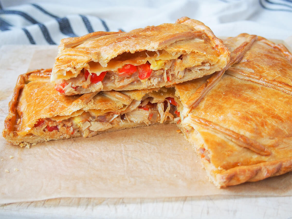 slice of empanada gallega sitting on top of rest of pie, showing filling in slice and golden pastry on top of pie.