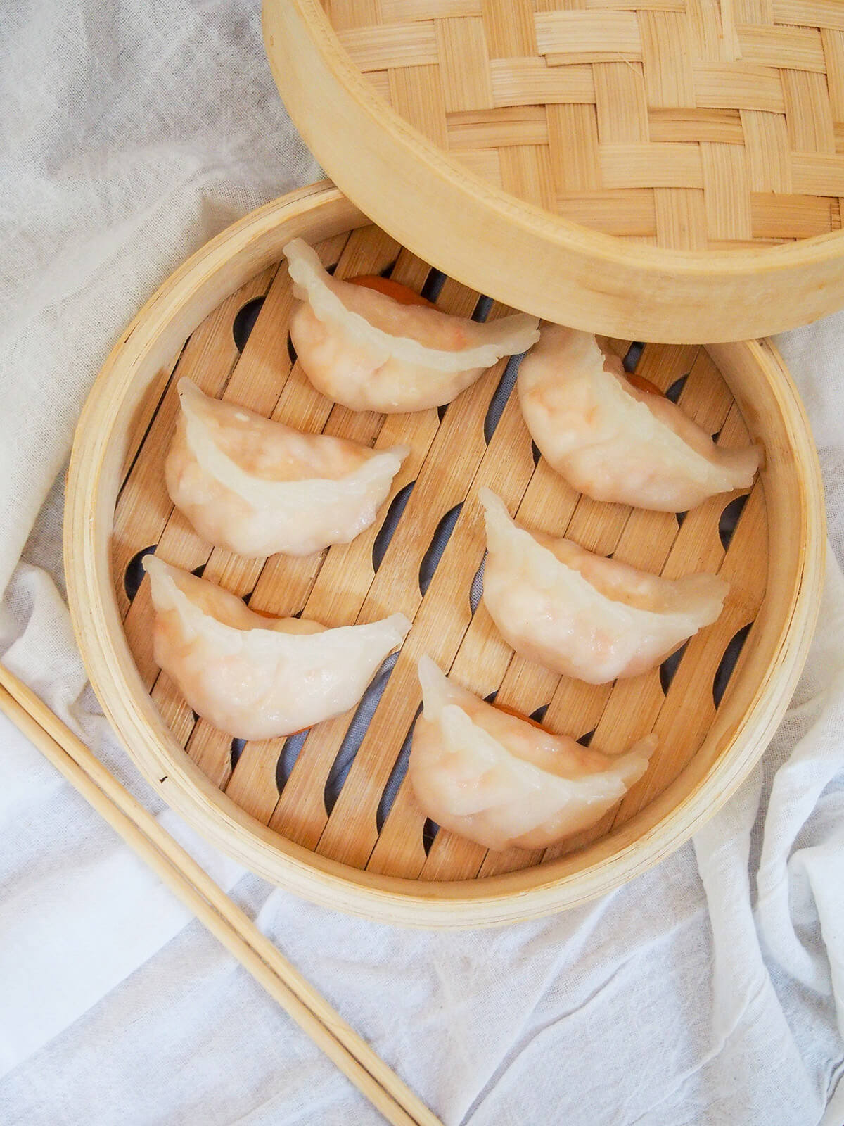 Har gow are a classic and delicious Cantonese dim sum, often called crystal shrimp dumplings due to their slightly translucent wrapper. The filling is simple as it's all about the juicy shrimp. They can be fiddly at first, but you get in the flow and it's well worth the effort. overhead view of har gow crystal shrimp dumplings in bamboo steamer with lid resting on one edge and chopsticks below