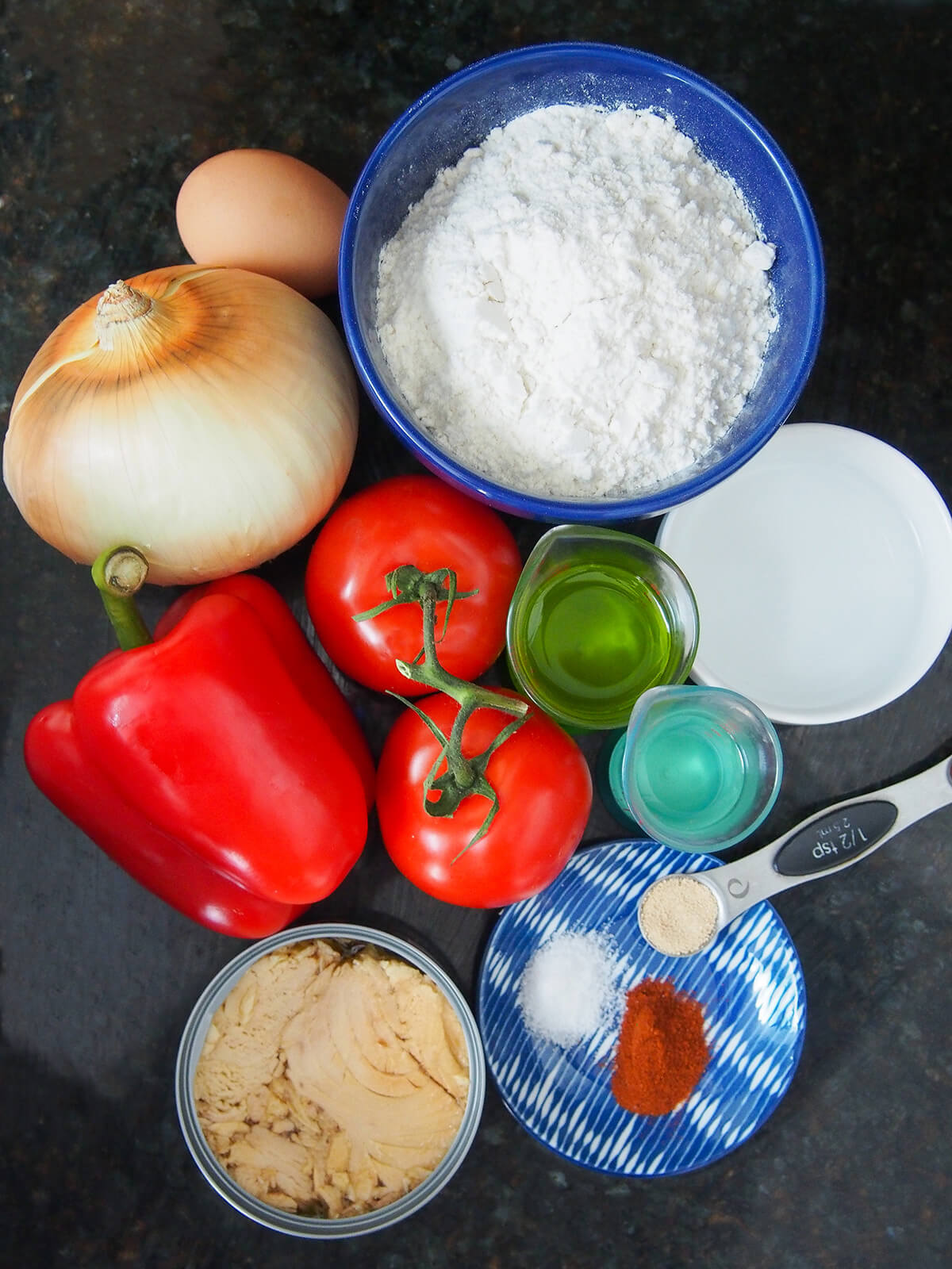 bowl of flour with measures of oil, wine, can of tuna, pepper, onion, tomatoes and egg around.