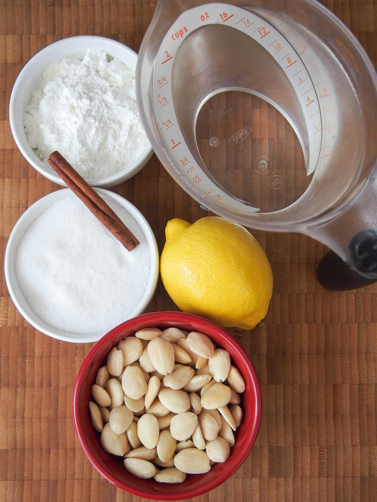 bowls with almonds, sugar, corn starch and water with lemon next to them and cinnamon stick balanced between bowls.