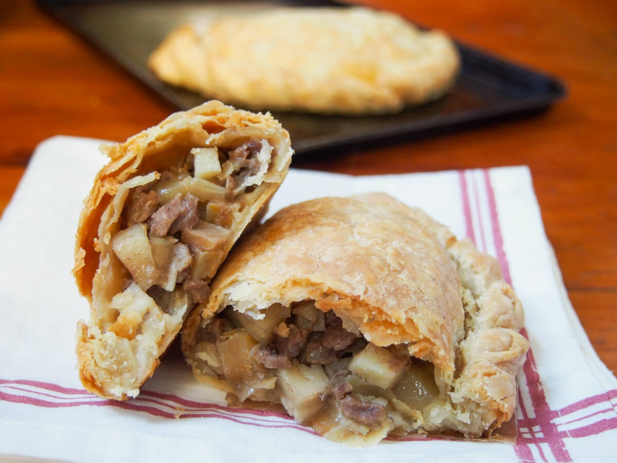 Cornish pasty cut open on top of kitchen towel with another on baking sheet blurred behind.