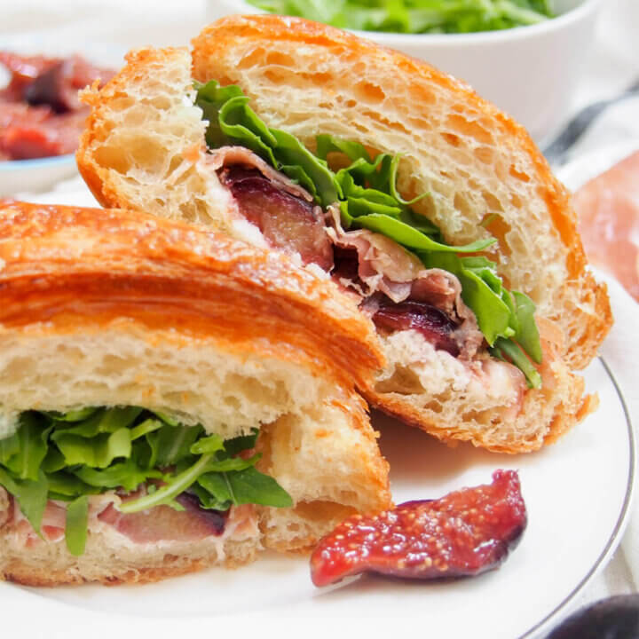 half of prosciutto and goat cheese croissant sandwich resting behind other half on white plate.