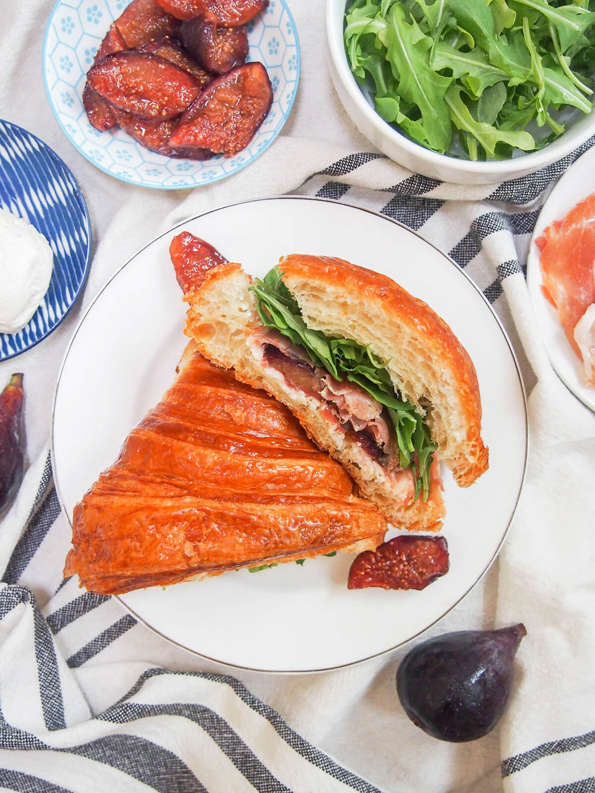 overhead view of half of prosciutto and goat cheese croissant sandwich on end showing filling, other half flat with dish of balsamic figs above.