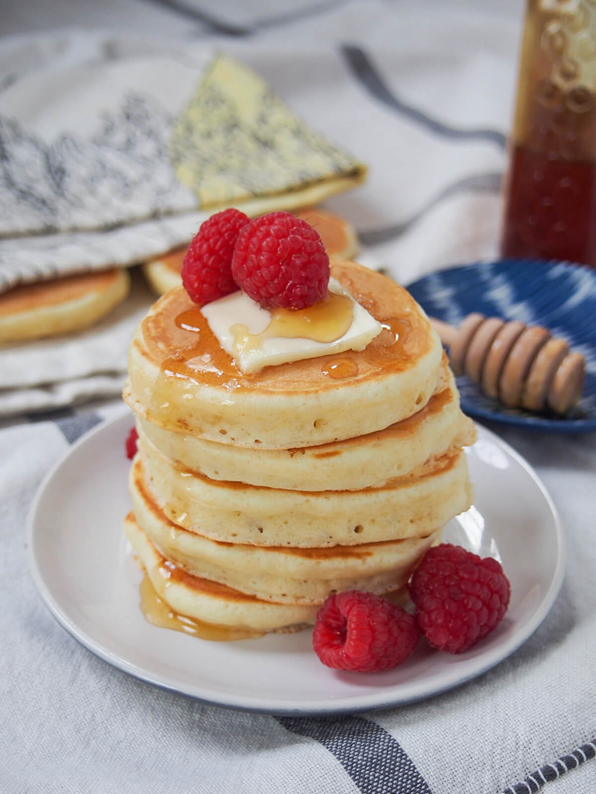 stack of Scotch pancakes on plate with honey mallet behind plate and cloth partly covering other pancakes.