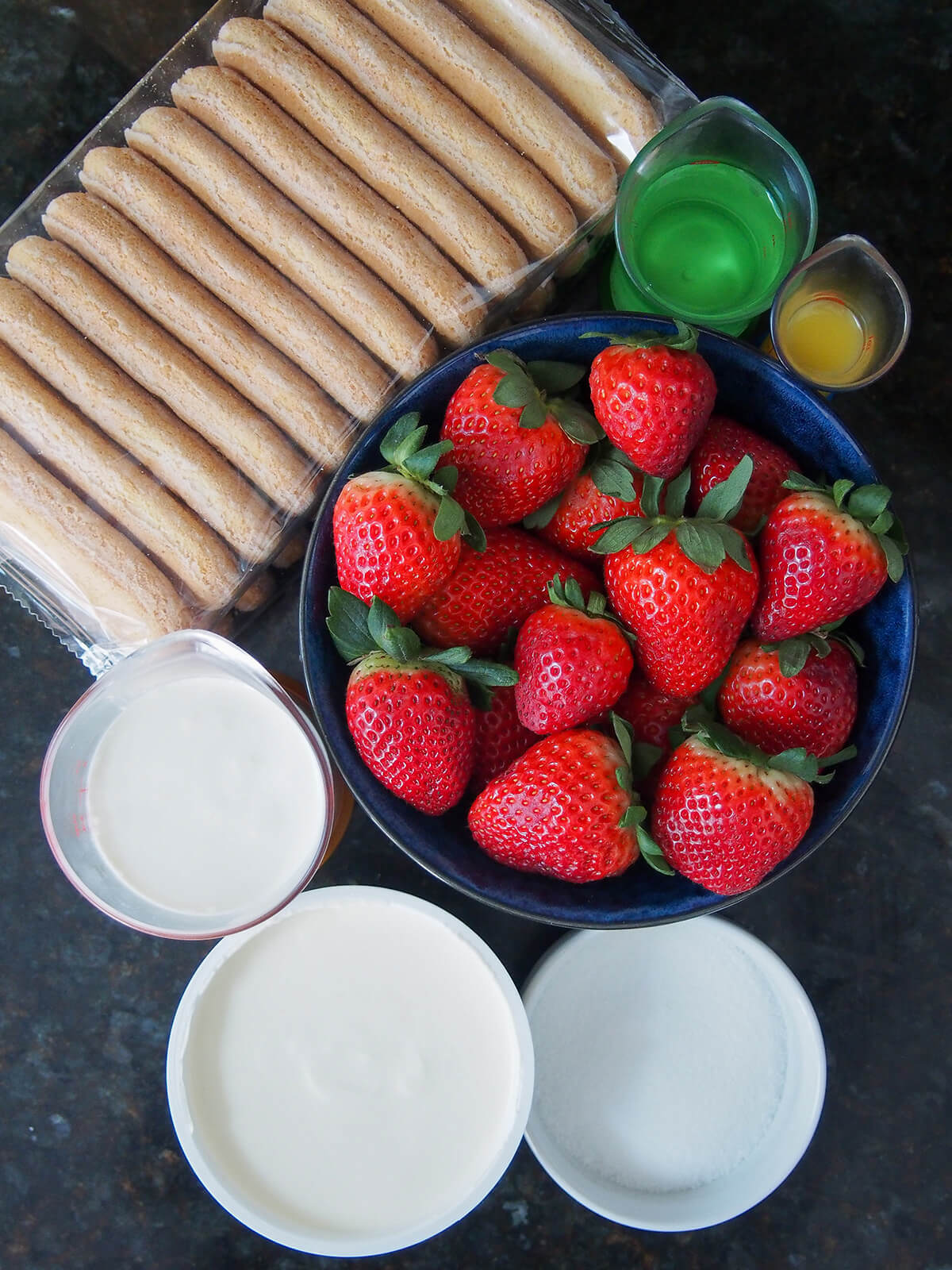 bowl of strawberries, container of mascarpone, measures with cream, water, packet of ladyfinger cookies and bowl of sugar from overhead.