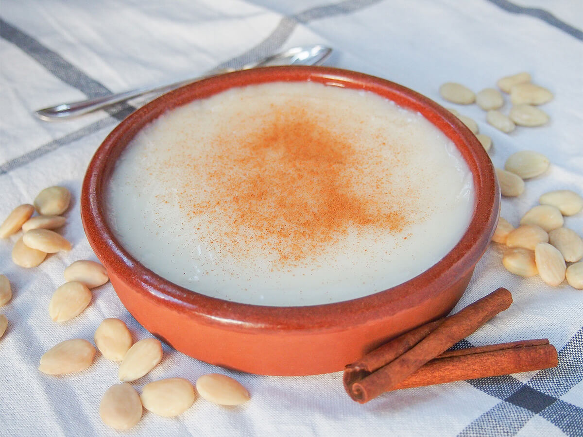 terracotta dish of menjarblanc almond pudding with cinnamon sticks in front and almonds either side.