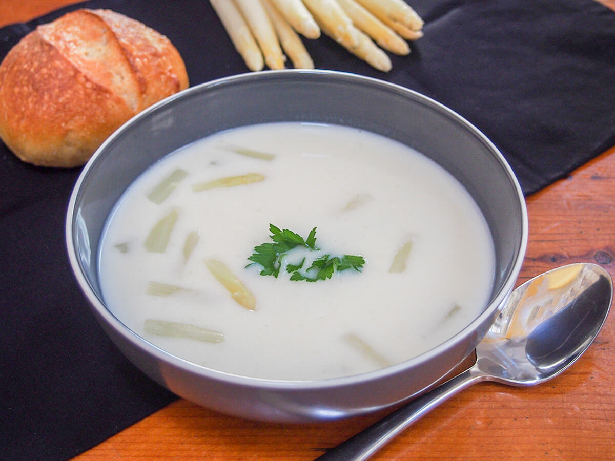 bowl of white asparagus soup with spoon to side of bowl and roll behind.