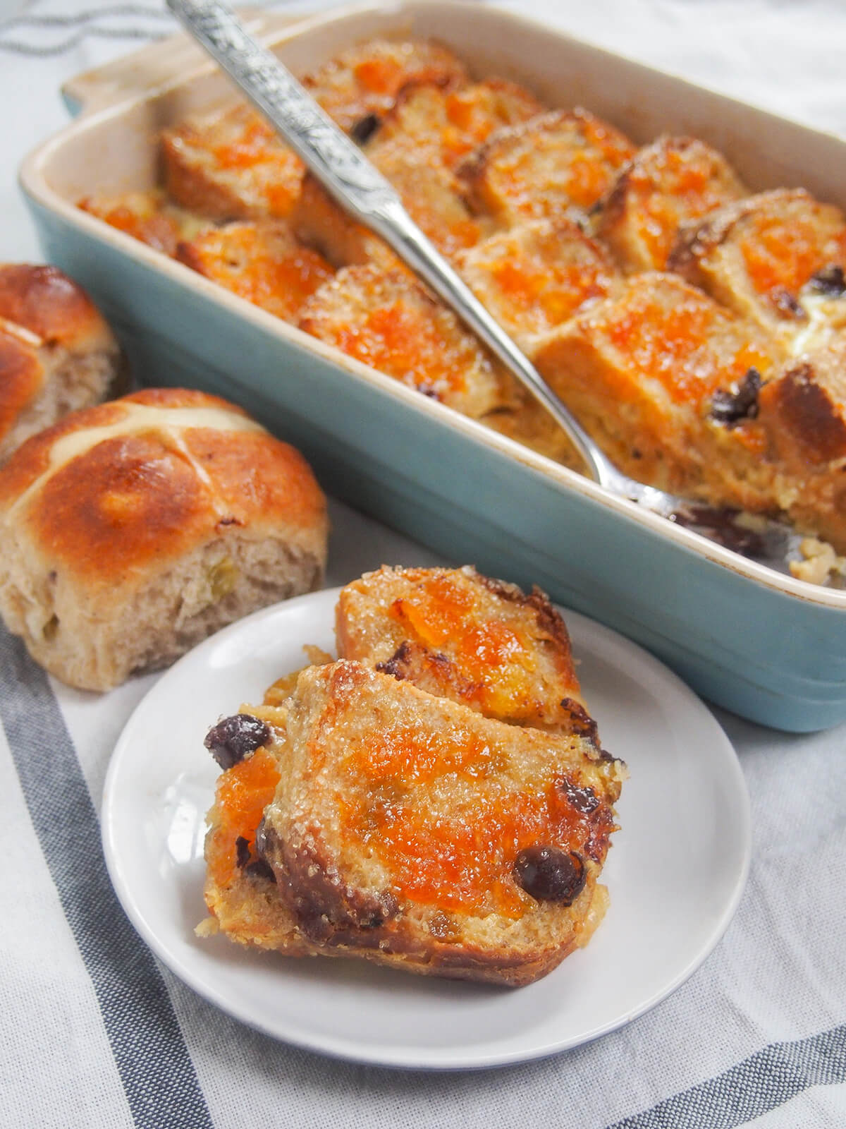 some of hot cross bun bread and butter pudding on small plate in front of dish with rest and spoon in dish.