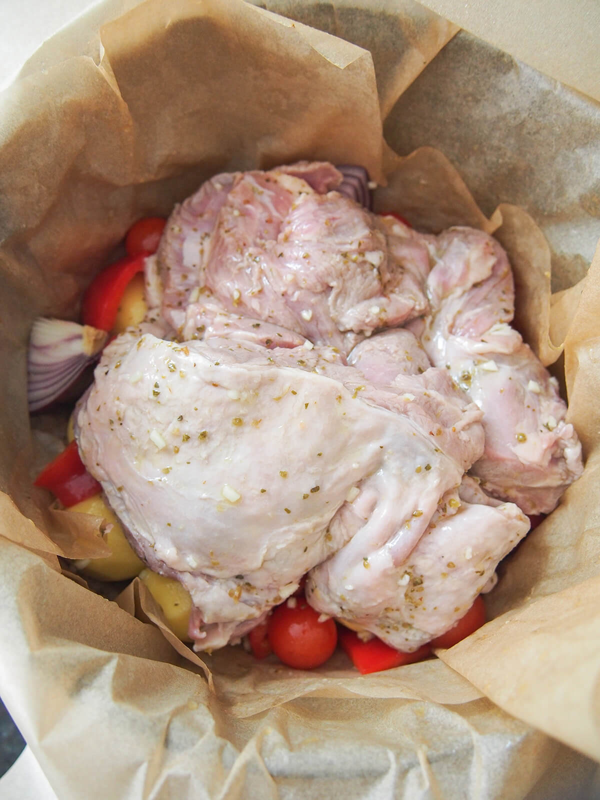 lamb and vegetables in parchment before closing and cooking piece of marinated lamb shoulder on top of potatoes, onion, pepper and tomatoes all sitting on pieces of parchment paper.