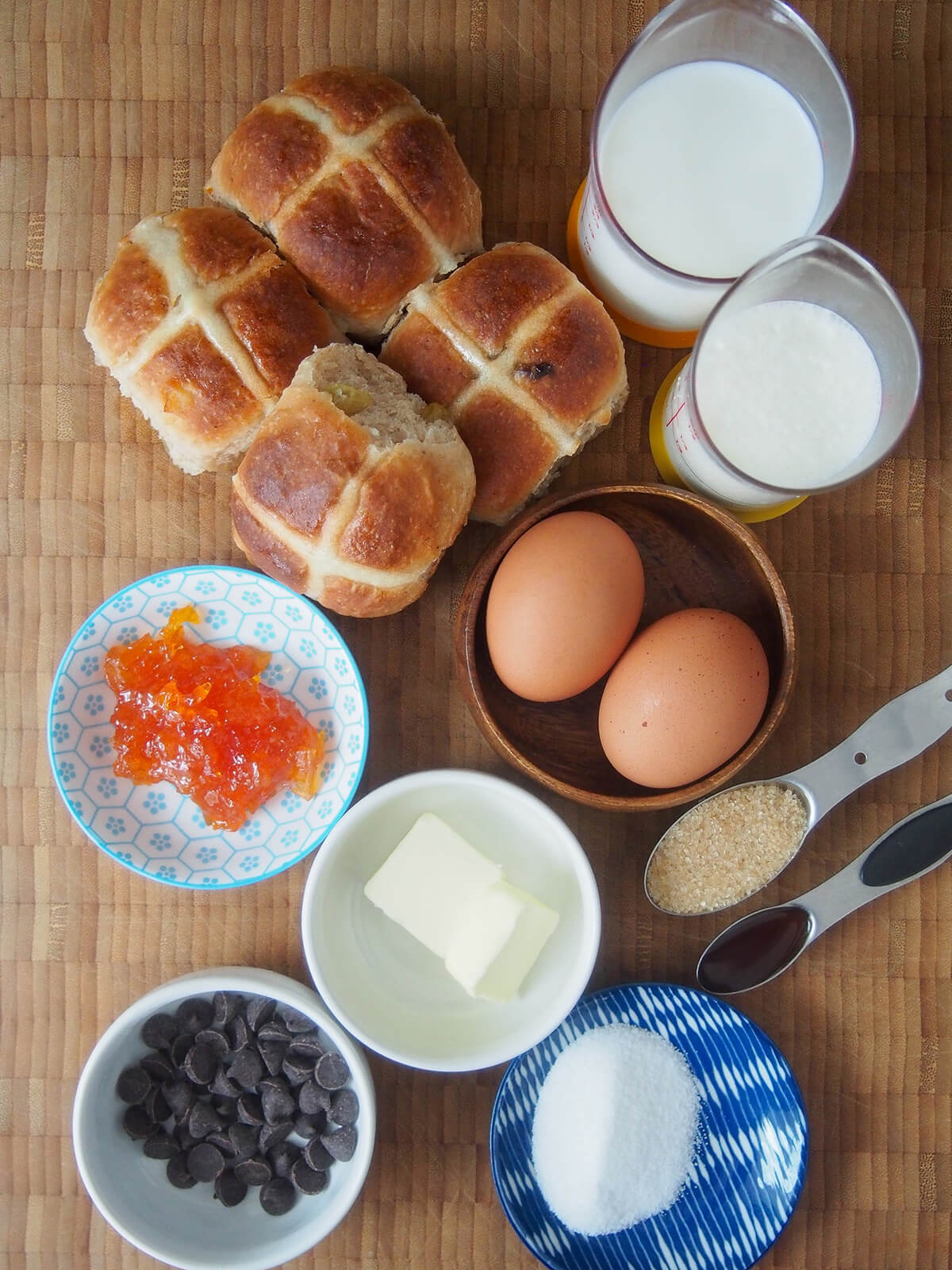 hot cross buns, measures with milk and cream, eggs in a dish, sugar, butter, chocolate chips and marmalade each in small dishes.