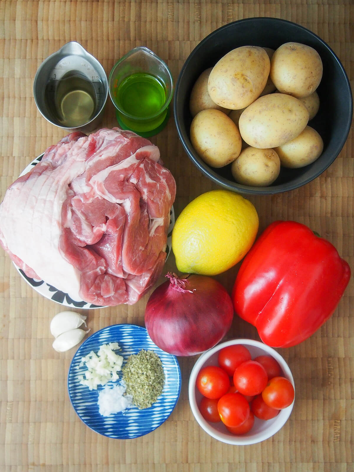 ingredients for lamb kleftiko bowl of baby potatoes, pepper, lemon, red onion, bowl of cherry tomatoes, small plate with herbs and garlic, piece of lamb and measures of oil and wine.