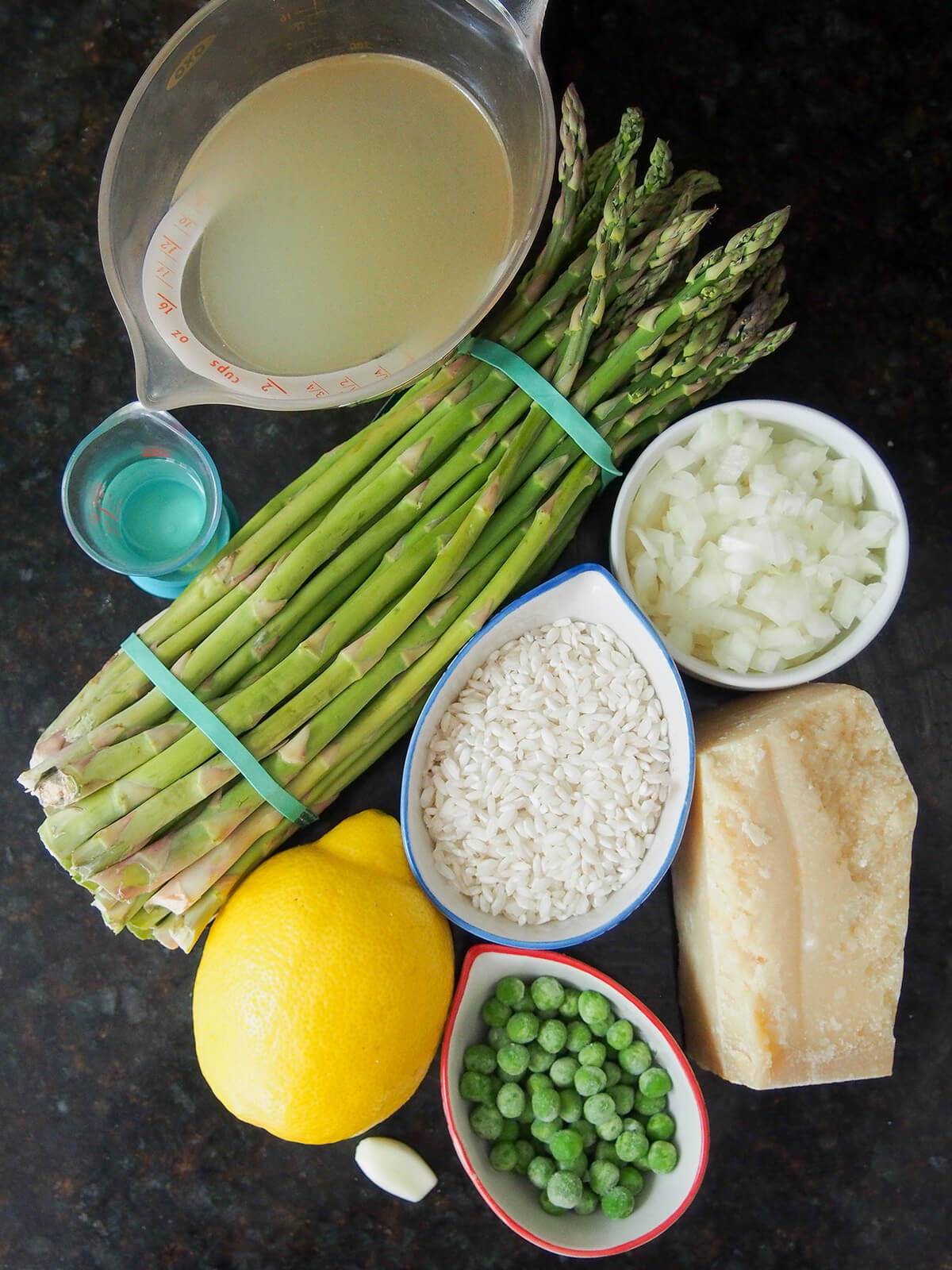 measure with stock, bunch of asparagus, bowls with rice, onion, peas as well as lemon and block of parmesan.