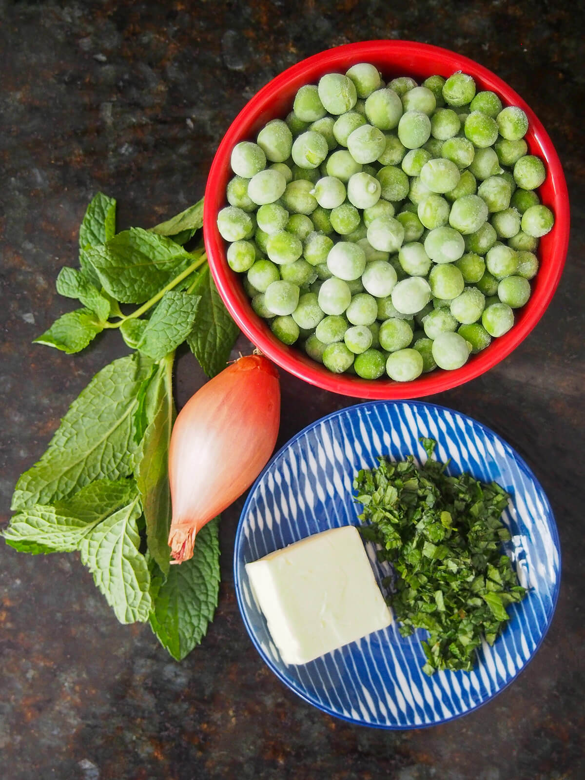 bowl of frozen peas, plate with chopped mint and piece of butter, shallot and bunch of mint to side.