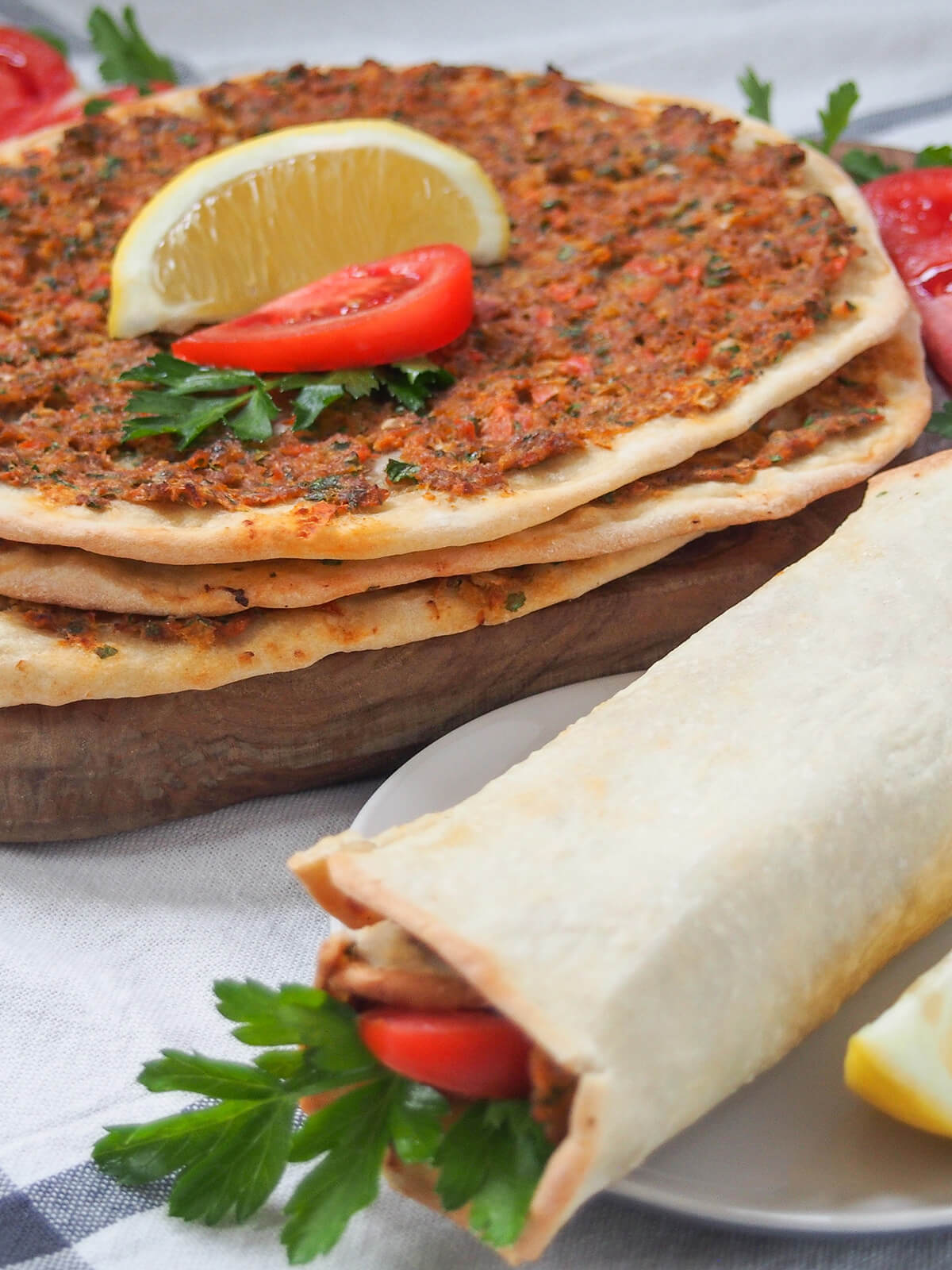 stack of lahmacun Turkish pizza towards back with lemon wedge on top with a rolled pizza in foreground.
