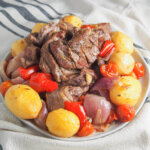lamb kleftiko on plate with potatoes, tomatoes and pepper around the lamb.