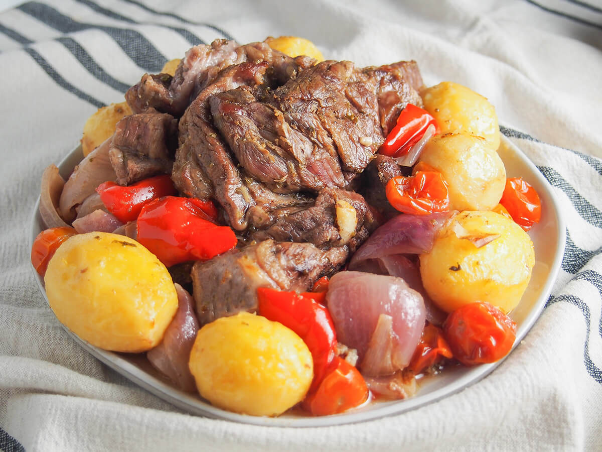 side view of plate of lamb kleftiko with potatoes, tomatoes, pepper and onion around the lamb