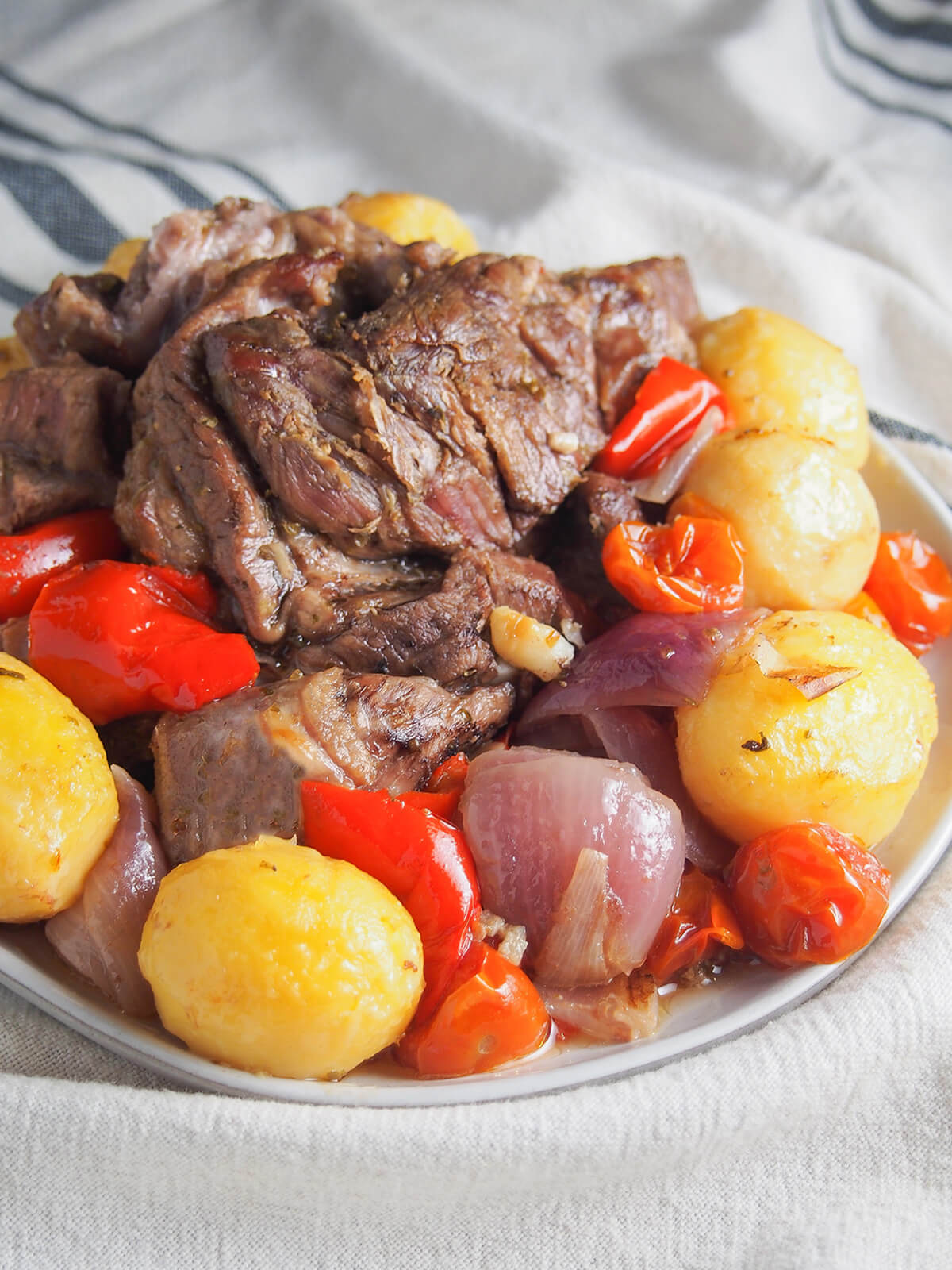 Lamb kleftiko is a wonderfully tasty Greek slow roasted lamb dish. The lamb becomes so tender and the vegetables cooked in with the meat to take on the delicious flavors, too. You need a little patience, but not much effort. And it's well worth the wait. lamb kleftiko with potatoes, pepper, tomatoes and onion around lamb on plate.
