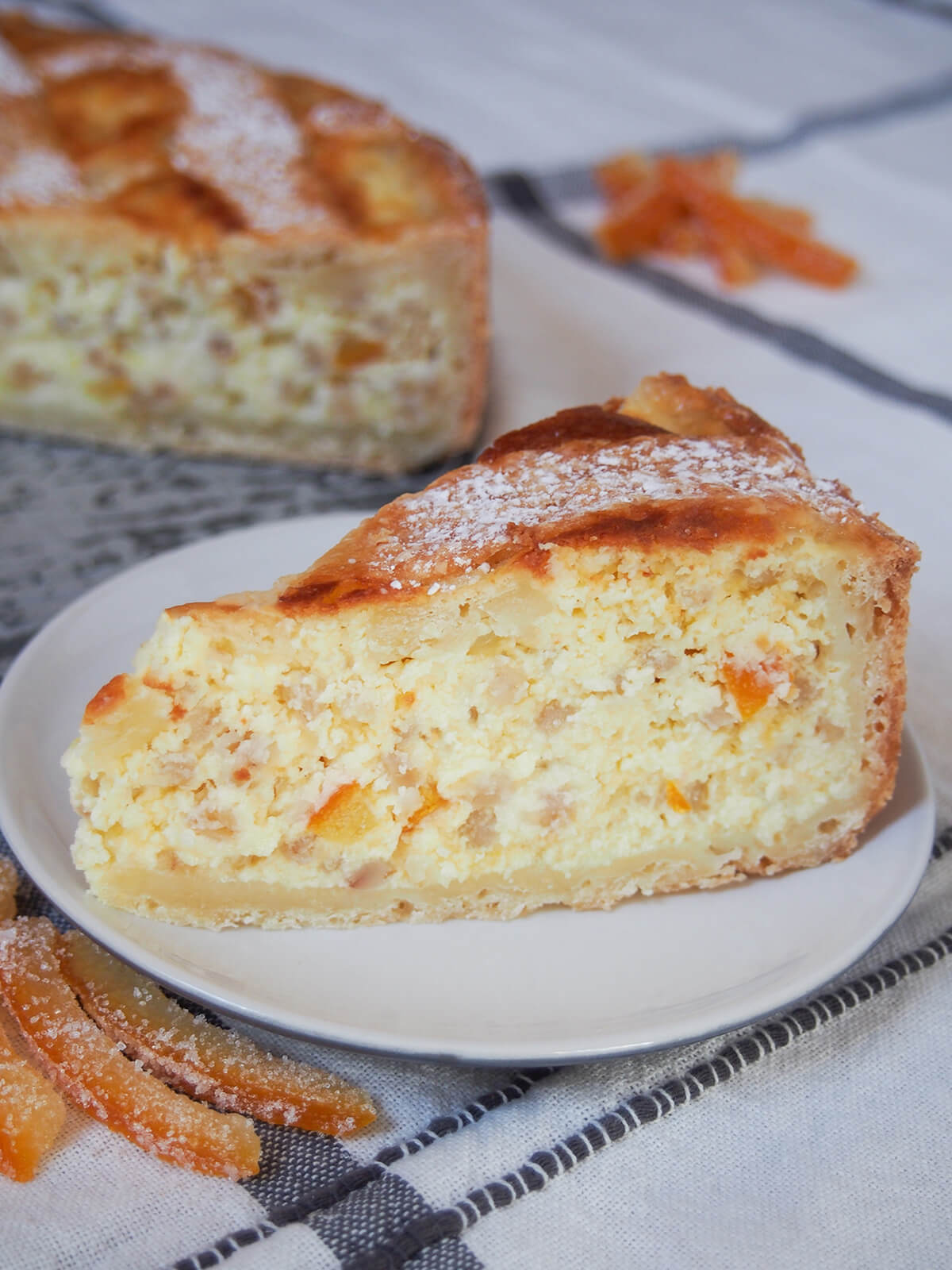 Pastiera napoletana is a lovely Italian tart/cake that's a traditional Easter dessert. It has a wonderfully creamy filling, gently aromatic with orange, and a crisp, buttery crust. A delicious treat whenever you enjoy it. slice of pastiera napoletana on plate with more of tart behind.