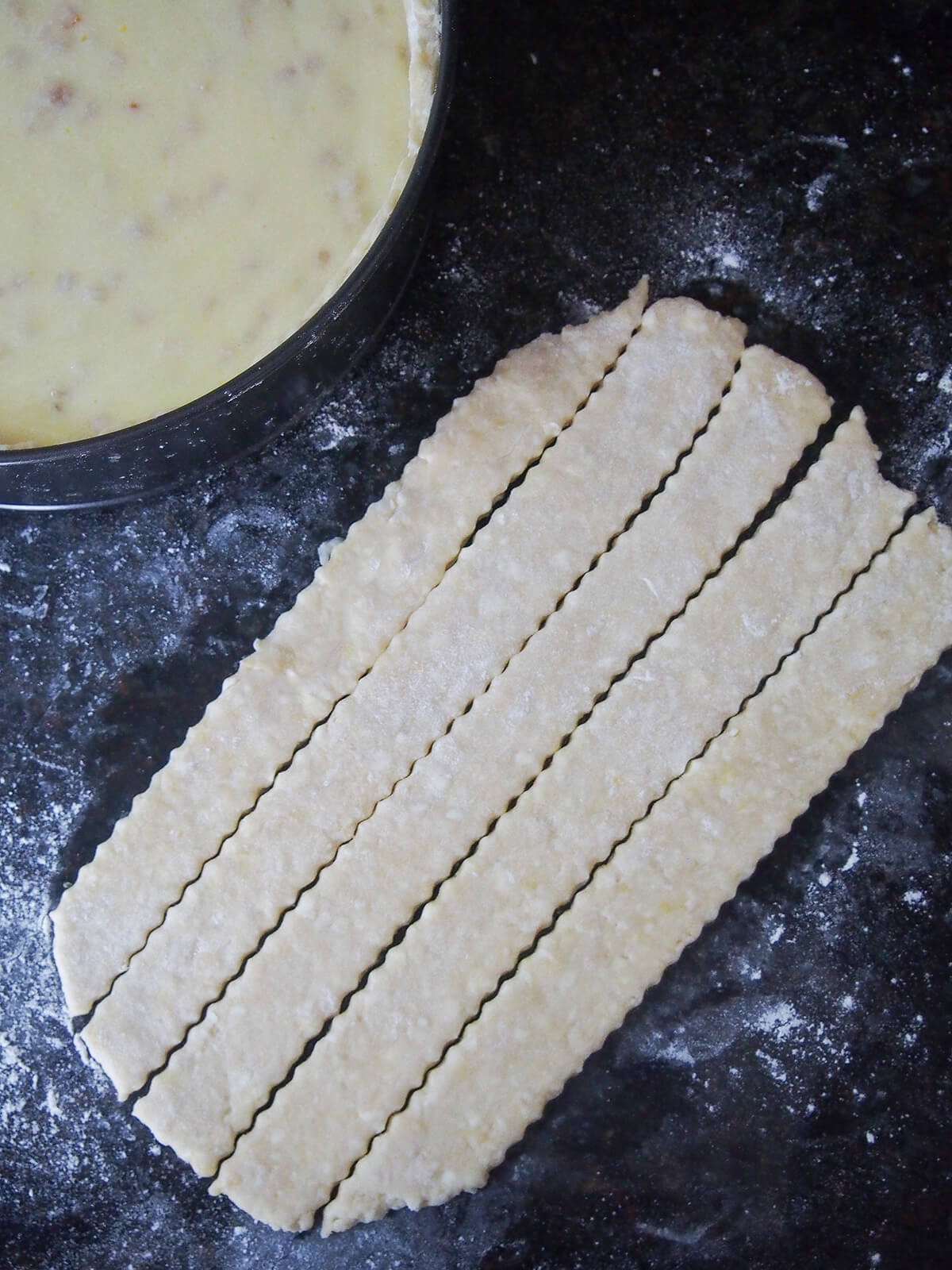 strips of pastry to top pastiera napoletana slices of pastry cut with zig ag edges next to baking tin with pastiera napoletana in it from overhead.
