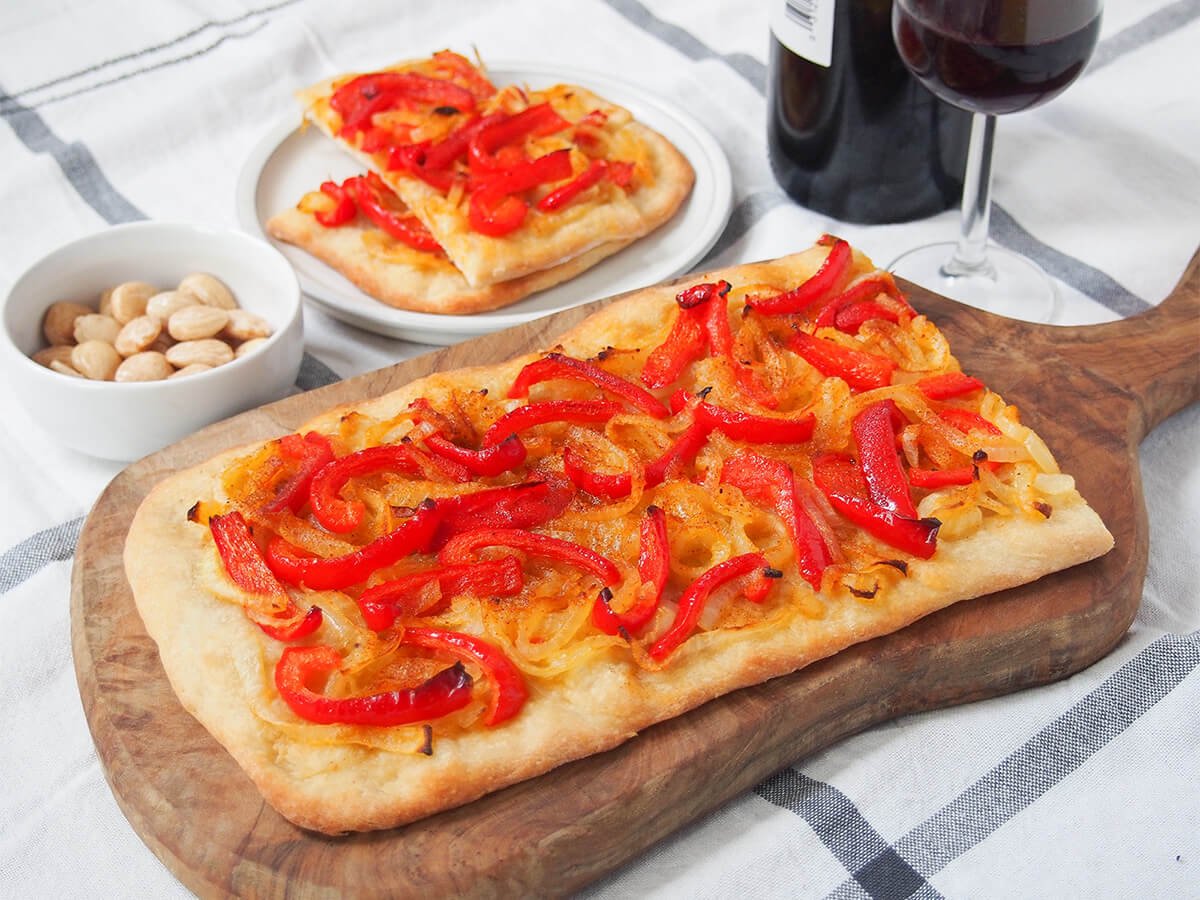 piece of Catalan coca on wooden board with slices on plate behind along with bowl of almonds and glass of wine.