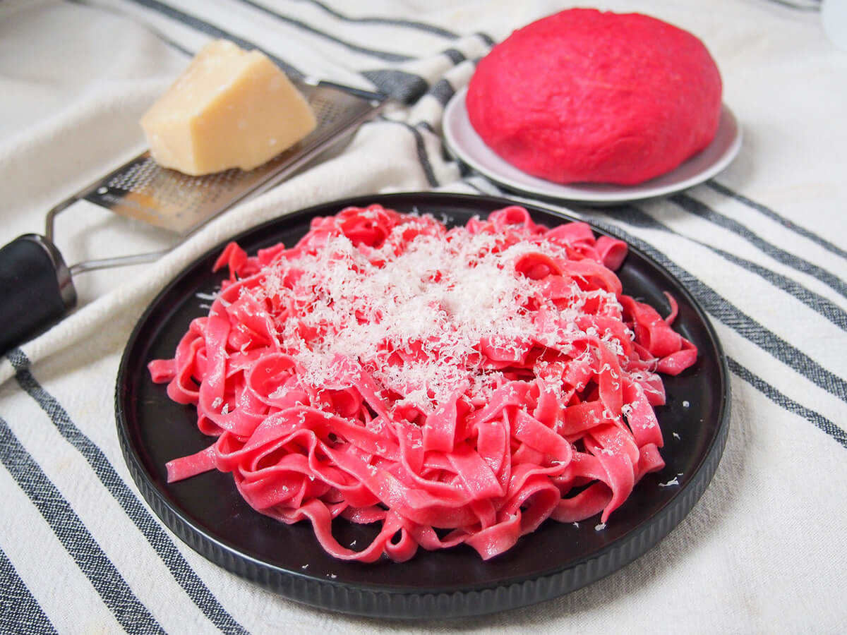 plate of beet pasta topped with parmesan with block of cheese and beet pasta dough behind.
