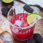 side view of glass of blackberry margarita with lime wedge and blackberry on stick on top of glass.