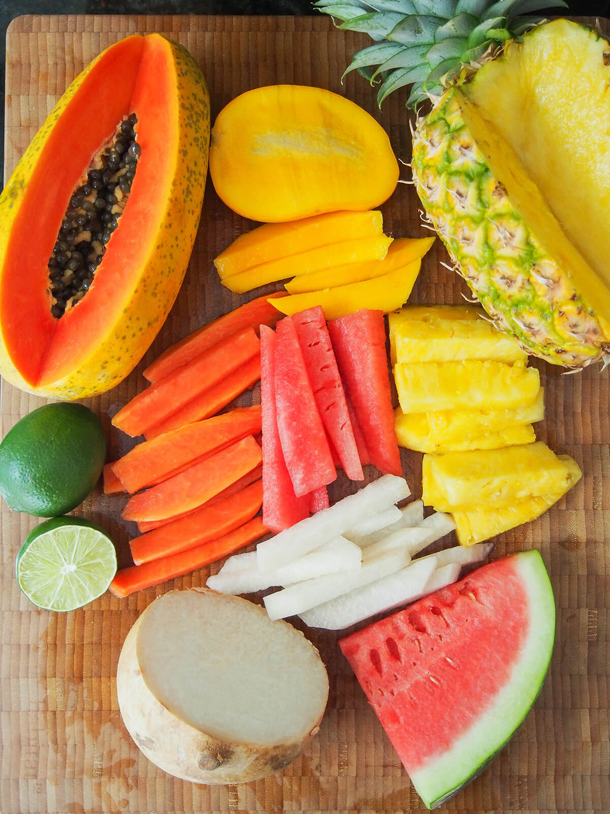 spears of cut pineapple, mango, jicama, watermelon and papaya with larger pieces of each fruit and limes around on chopping board.