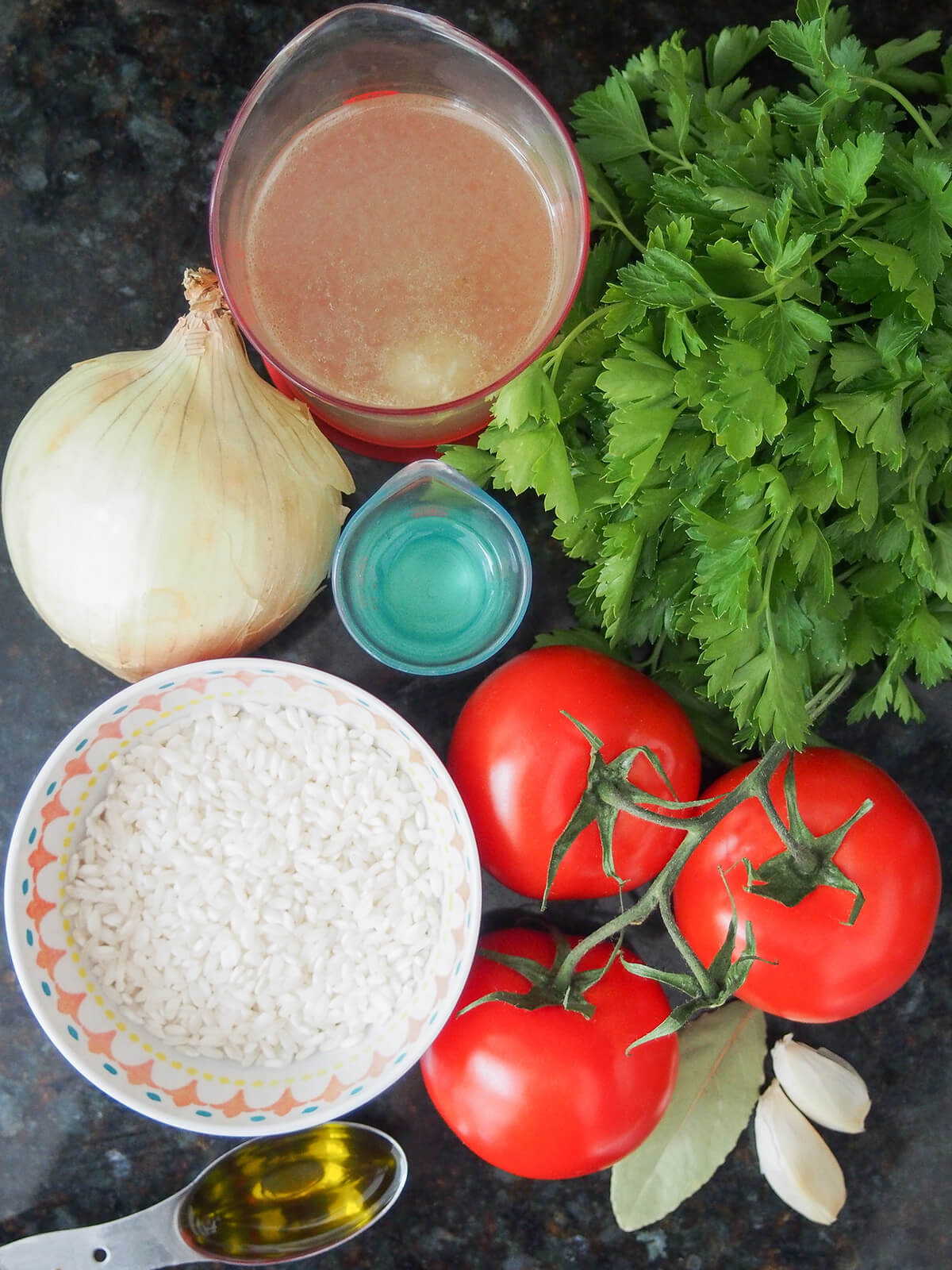 bowl of rice, bunch of tomatoes, onion, bunch of parsley with measures if stock, wine, garlic cloves and. bay leaf below.