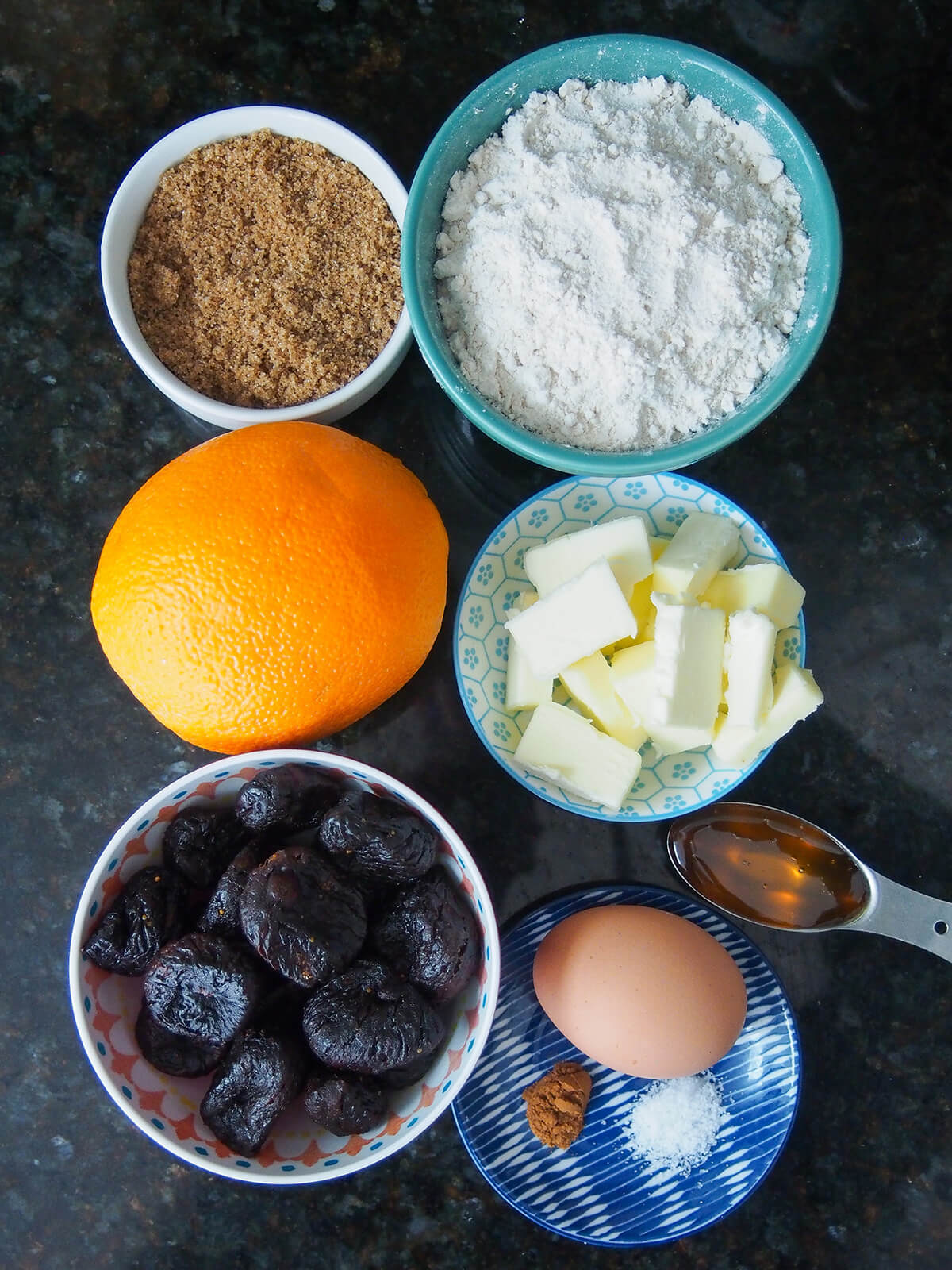 bowls of flour, sugar, butter, dried figs with orange, spoon of honey and plate with egg and salt in between.