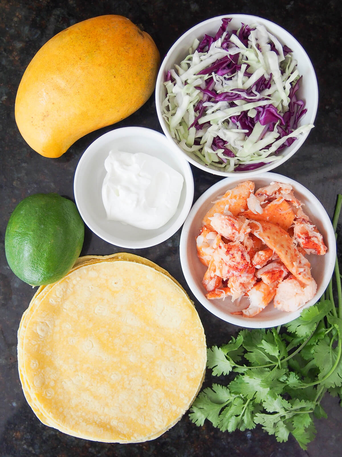 bowls of shredded cabbage, sour cream, lobster and stack of corn tortillas, mango, lime and some cilantro.