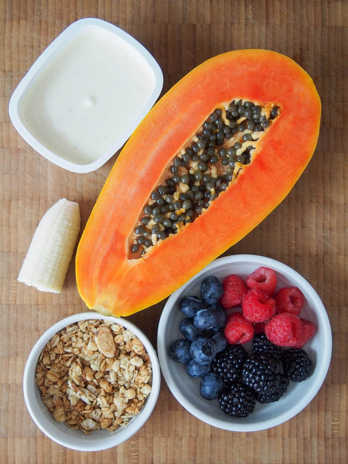 container of yogurt, half papaya, bowl of berries, bowl of granola and part banana on wooden board.