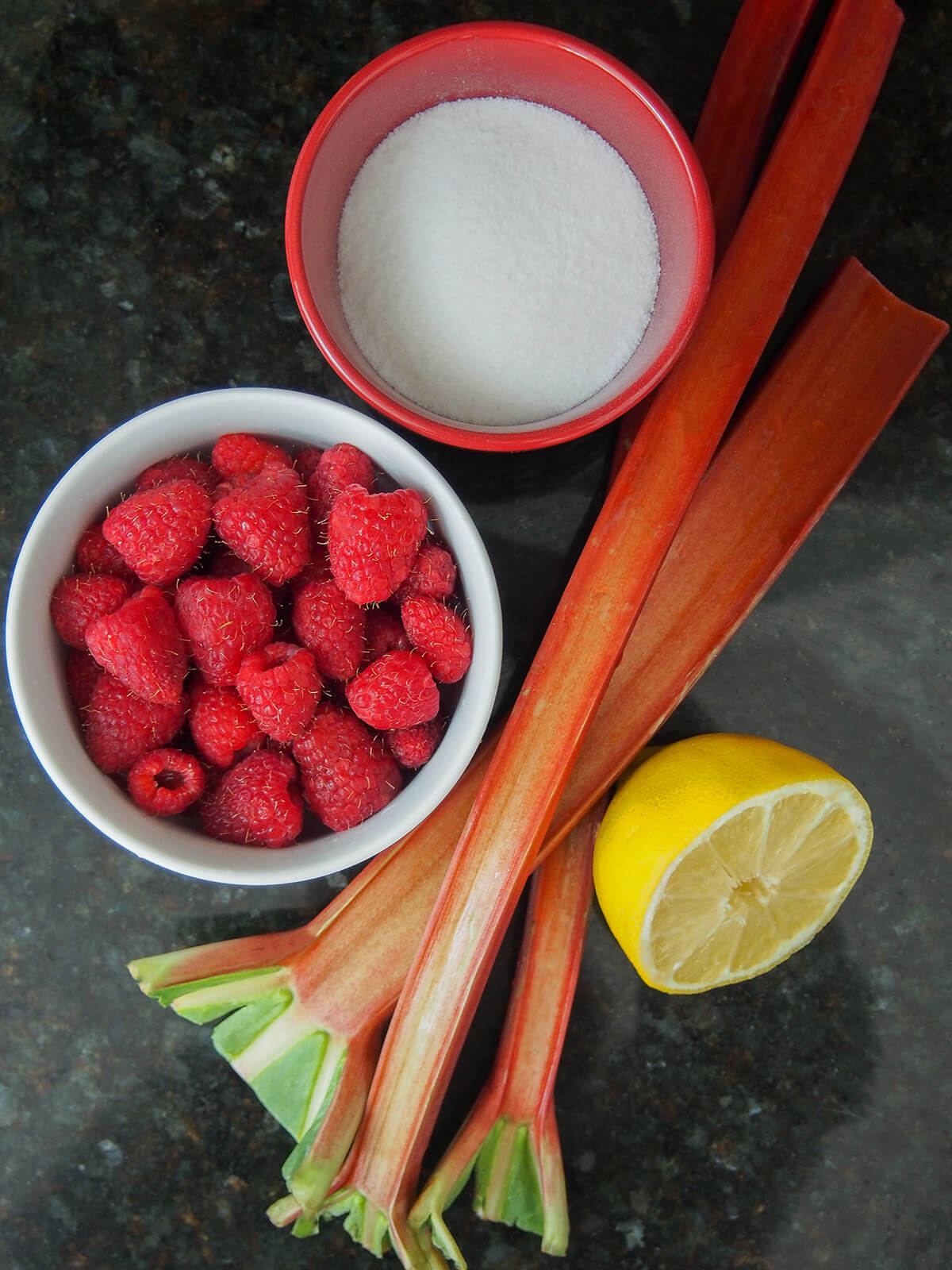 bowls of sugar, raspberries, part lemon with stalks of rhubarb in between.