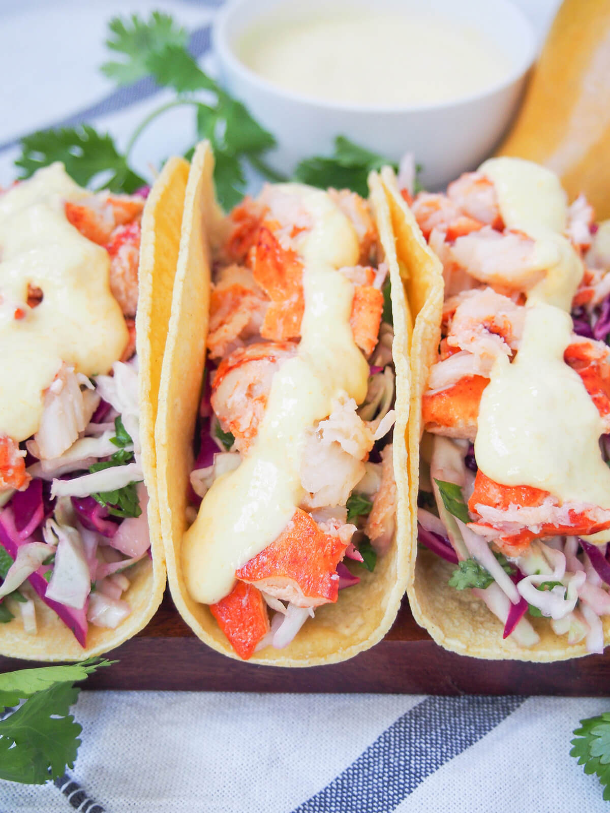 side view of three lobster tacos with bowl of mango crema in the background.