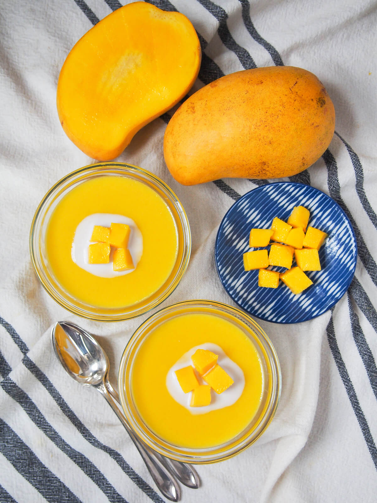 overhead view of two bowls of mango pudding with spoons to one side, mangoes above and small plate with mango chunks to side.