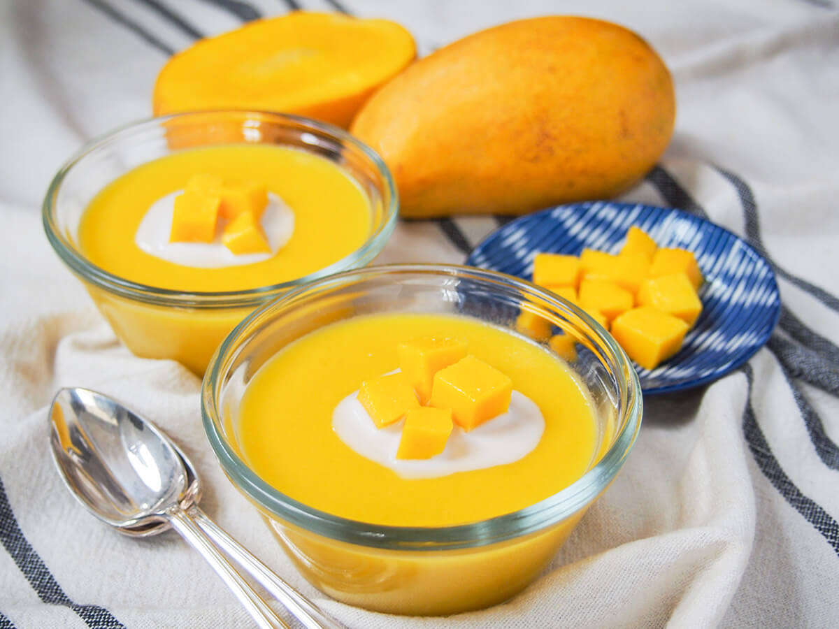 two bowls of mango pudding with mango, both whole and cut, behind bowls.