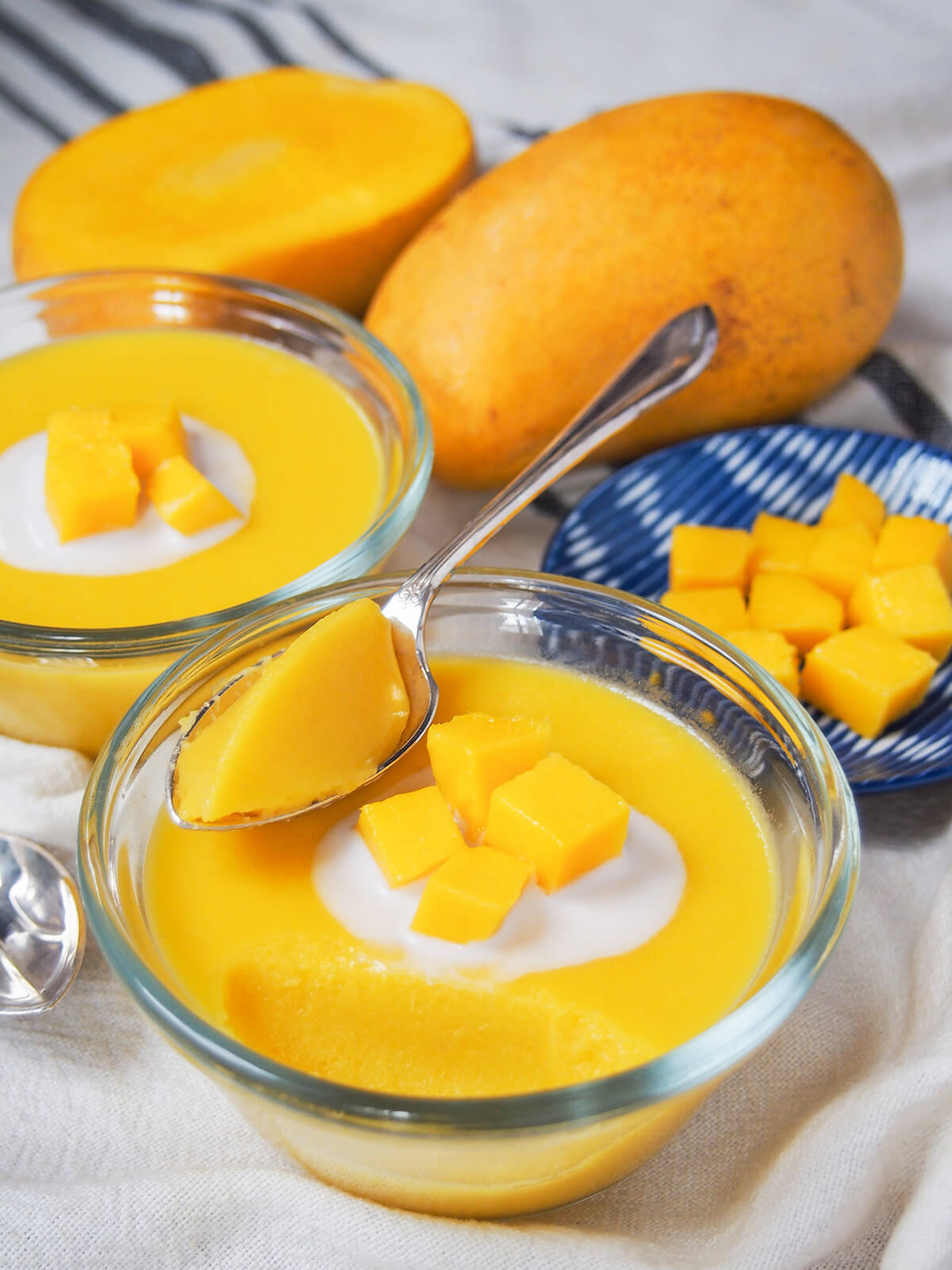 bowl of mango pudding with spoon sitting towards back and mango and part of other pudding behind.