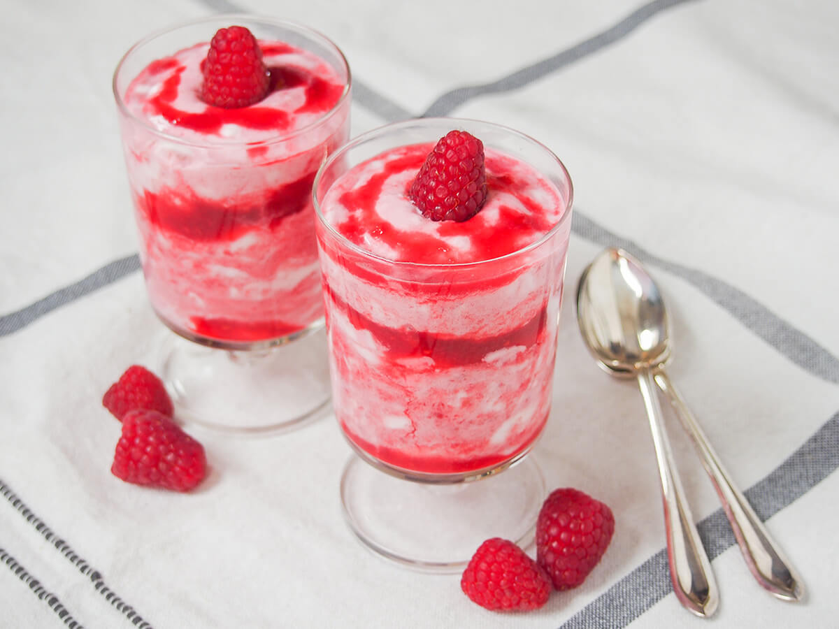 two glasses of raspberry fool with raspberries wither side of glasses and spoons to right.
