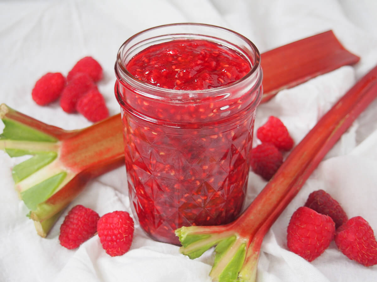 side view of raspberry rhubarb jam with stems of rhubarb and raspberries around jar.