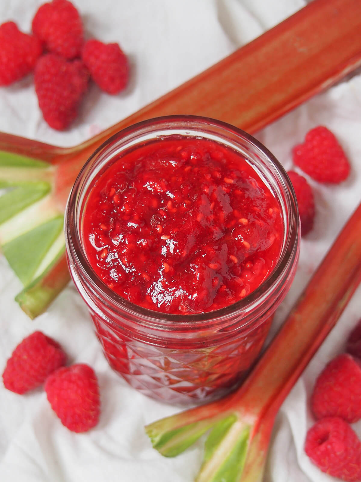 jar of raspberry rhubarb jam from overhead with rhubarb stalks and raspberries below jar.