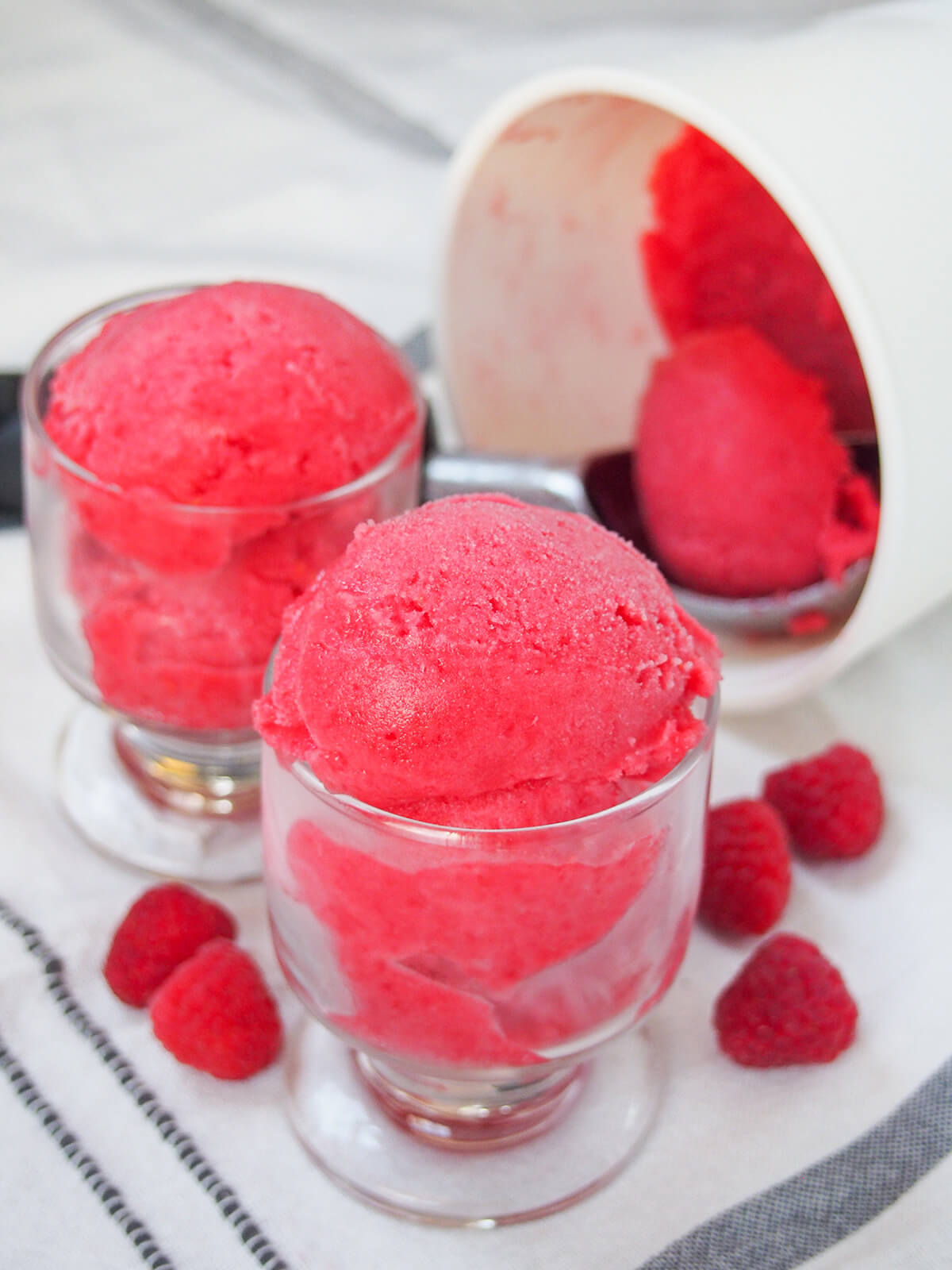 two glasses with raspberry sorbet with a scoop in side-on tub behind and raspberries around glasses.