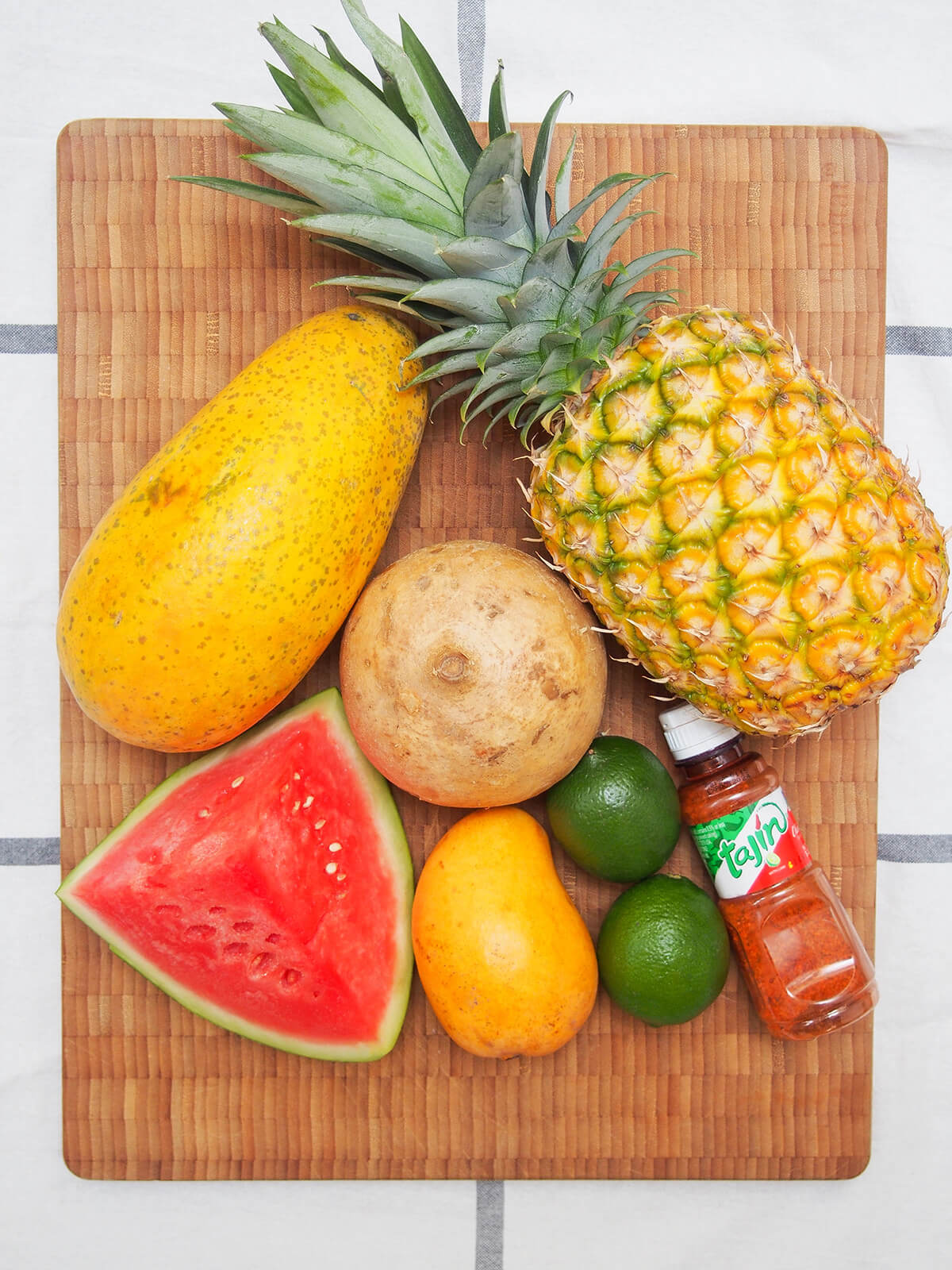 whole pineapple, jicama, papaya, mango and limes with chunk of watermelon and bottle of tajin to sides on wooden chopping board.