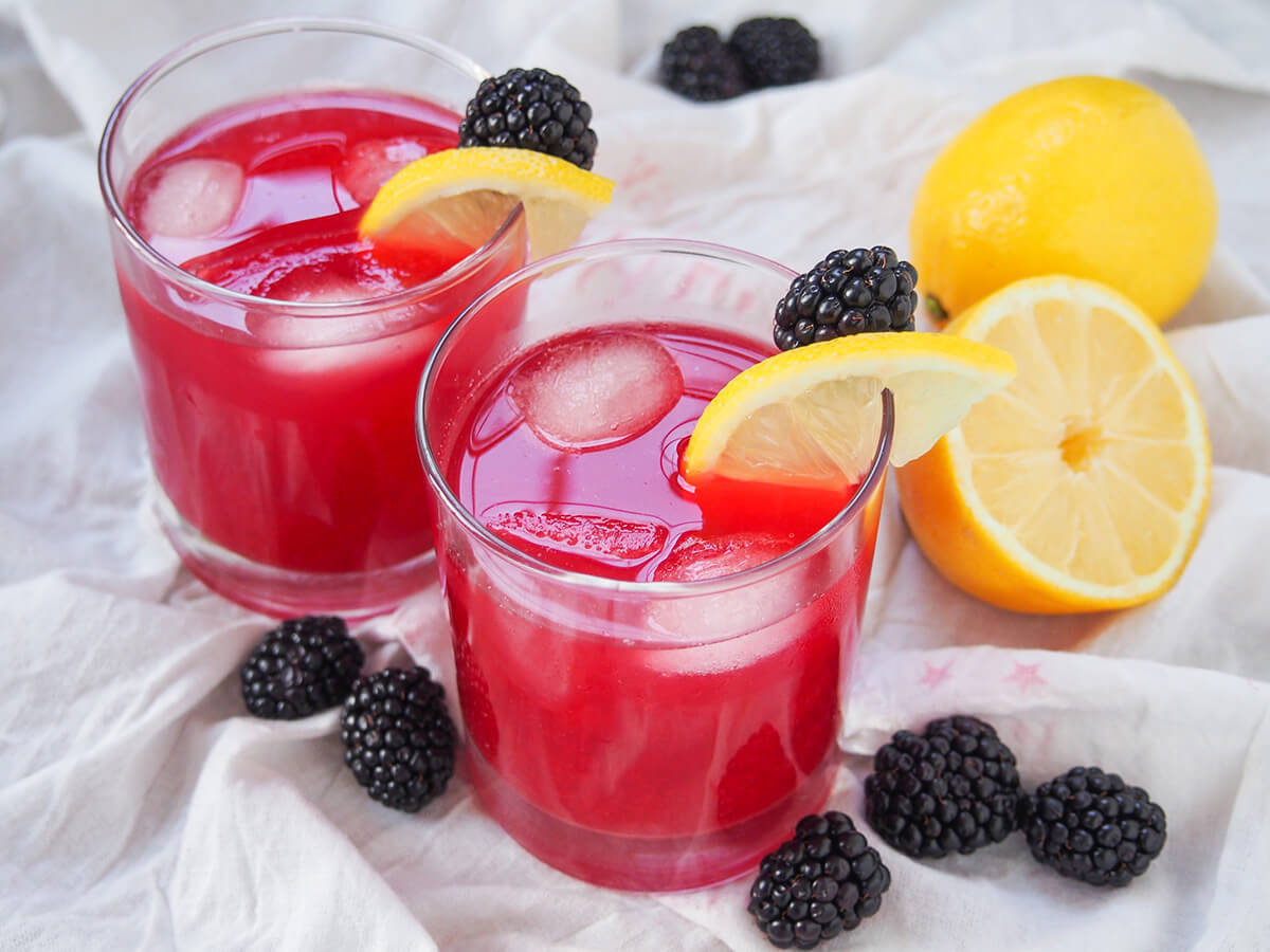 two glasses of blackberry lemonade with lemons behind and blackberries to side of glasses.