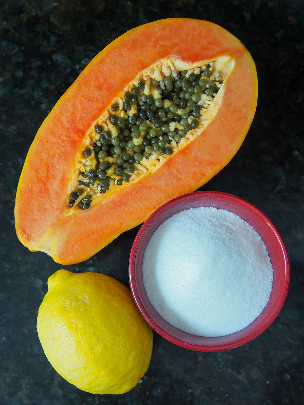 ingredients for papaya jam half papaya, bowl of sugar and a lemon viewed from overhead.