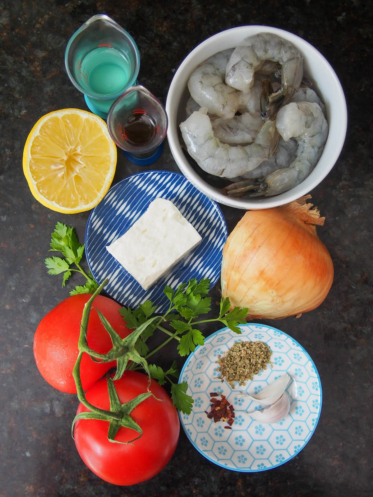 bowl of shrimp, plate with piece of feta on it, part onion, tomatoes, lemon and dish with garlic and herbs.