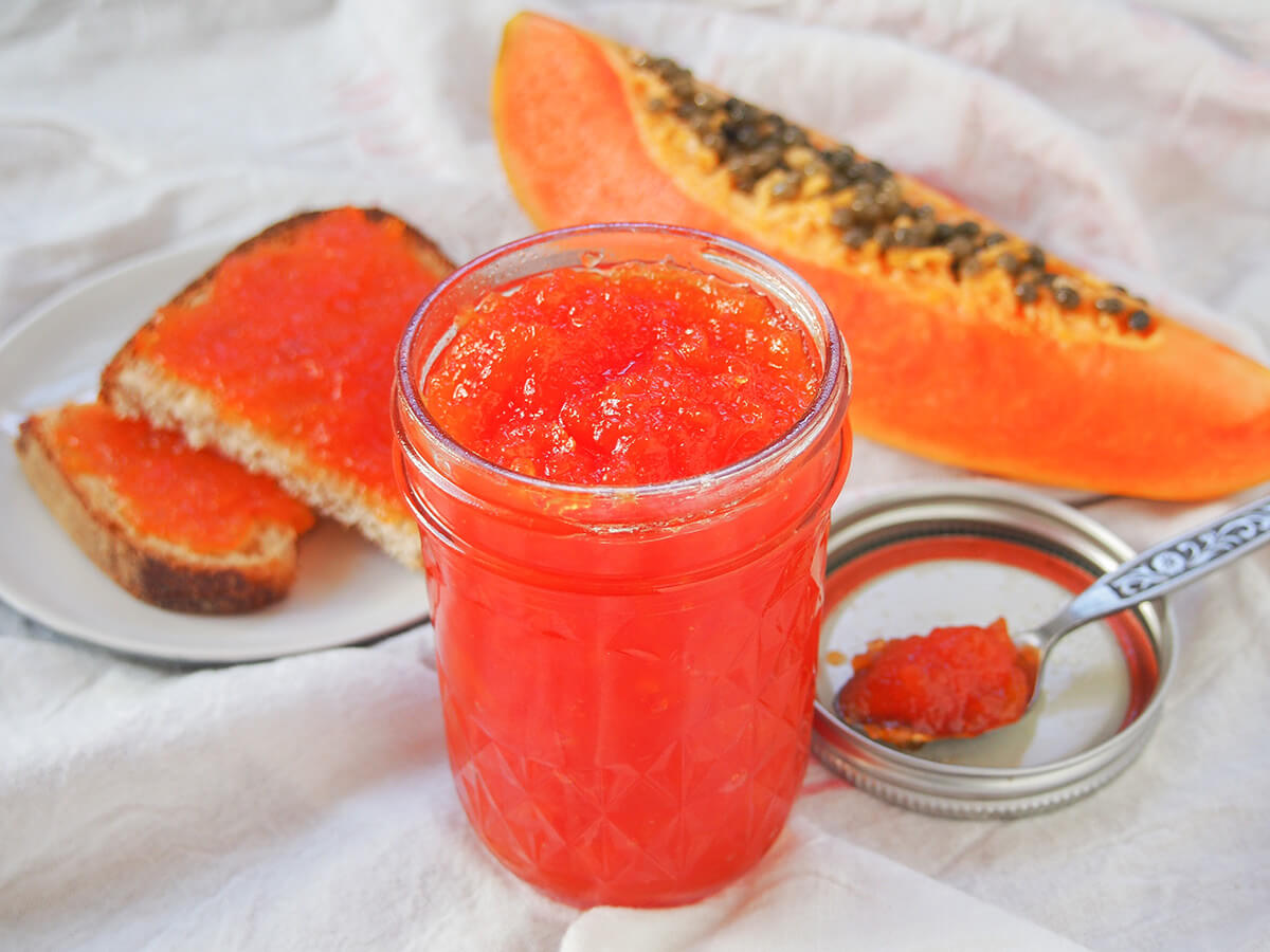 papaya jam jar with toast behind jar of papaya jam with slice of papaya and some on bread behind.