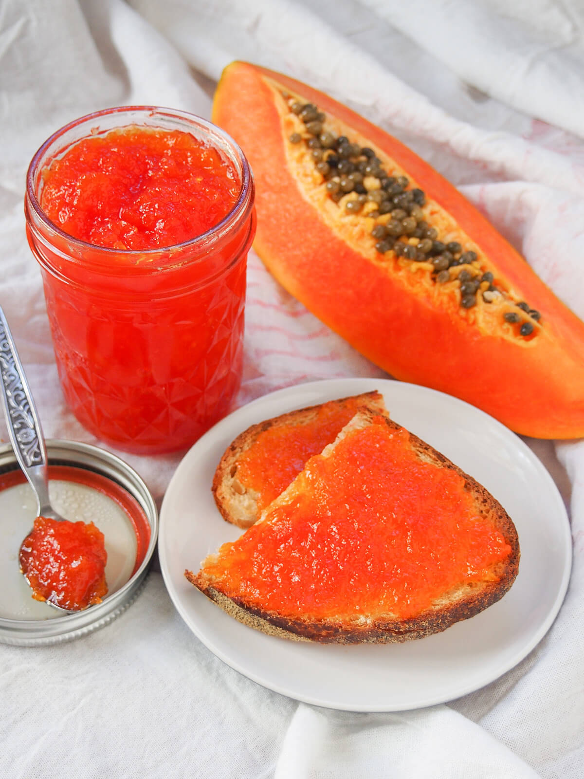 This papaya jam is easy to make, with a lovely tropical fruit flavor. It's gently aromatic and great both as a sweet spread and paired with cheese. pieces of bread topped with papaya jam with jar to side and piece of papaya behind plate.