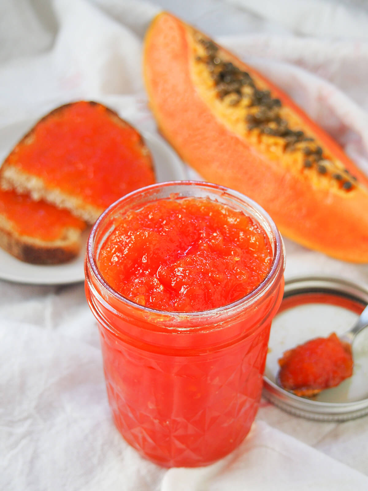 This papaya jam is easy to make, with a lovely tropical fruit flavor. It's gently aromatic and great both as a sweet spread and paired with cheese. jar of papaya jam with piece of papaya and toast with the jam on it slightly blurred in background.