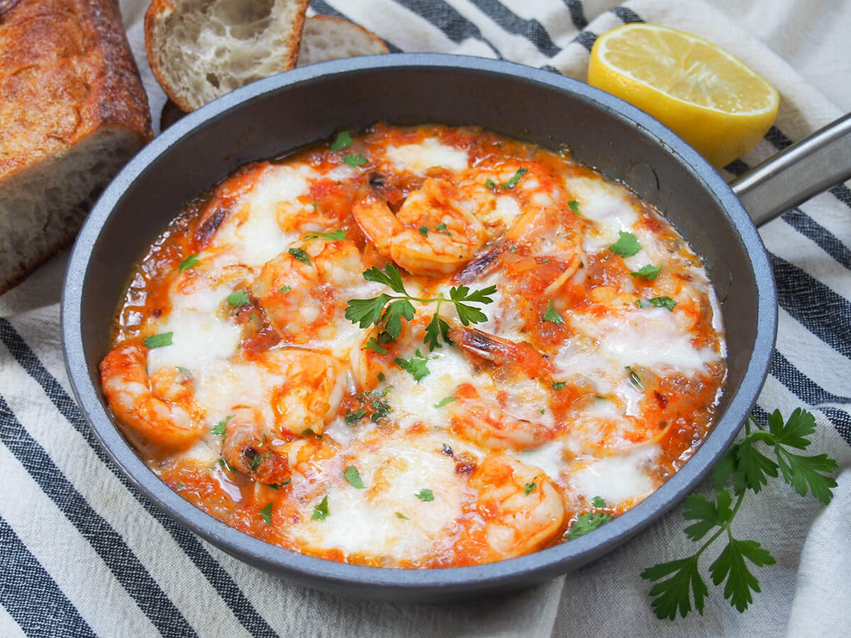 skillet of shrimp saganaki with bread and lemon behind.