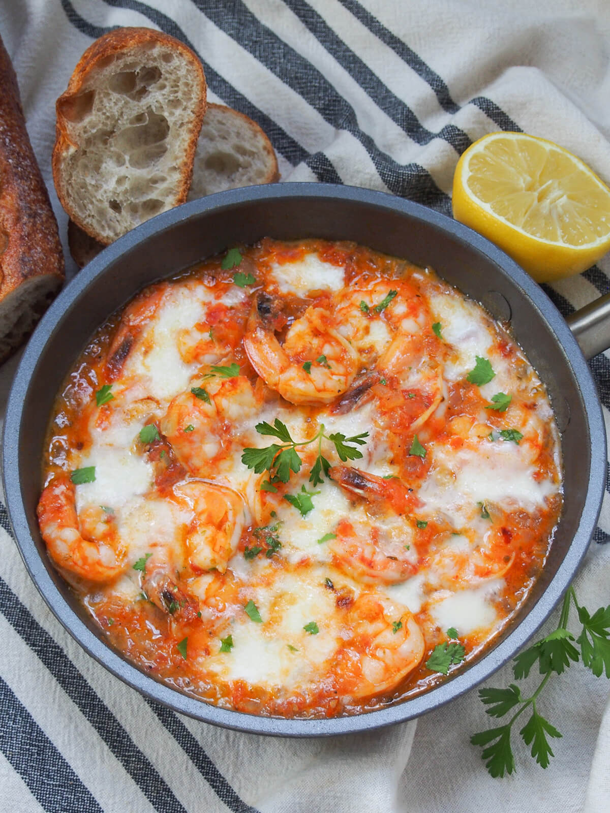 skillet of shrimp saganaki with slices of bread and a part lemon above and parsley sprig below.