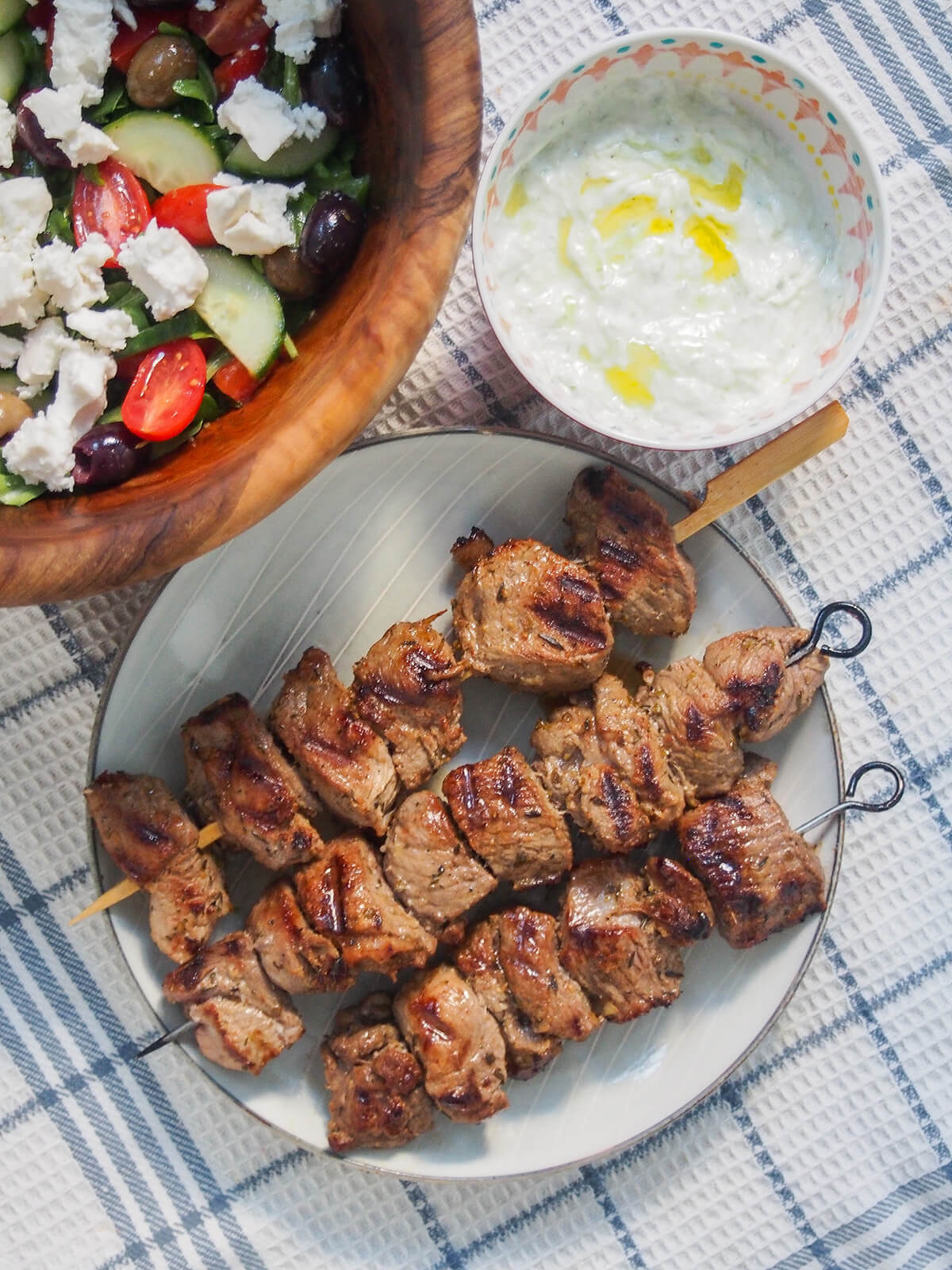 This flavorful Greek lamb marinade is easy to prepare and perfect to make lamb souvlaki skewers, or for larger chops or steaks. Delicious with pita, salad, tzatziki and more. overhead view of a plate of Greek lamb souvlaki with bowl of tzatziki and salad above.